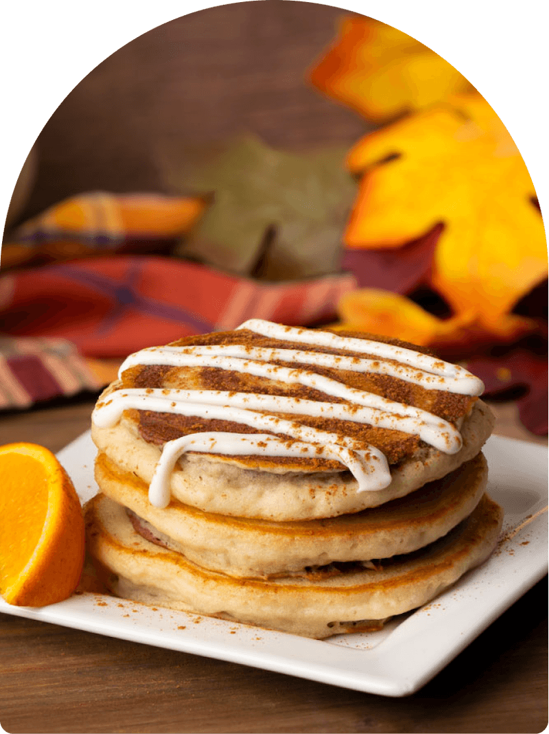Stack of cinnamon roll pancakes topped with cream cheese icing and cinnamon, served with a fresh orange slice on a white plate, with fall leaves and plaid fabric in the background