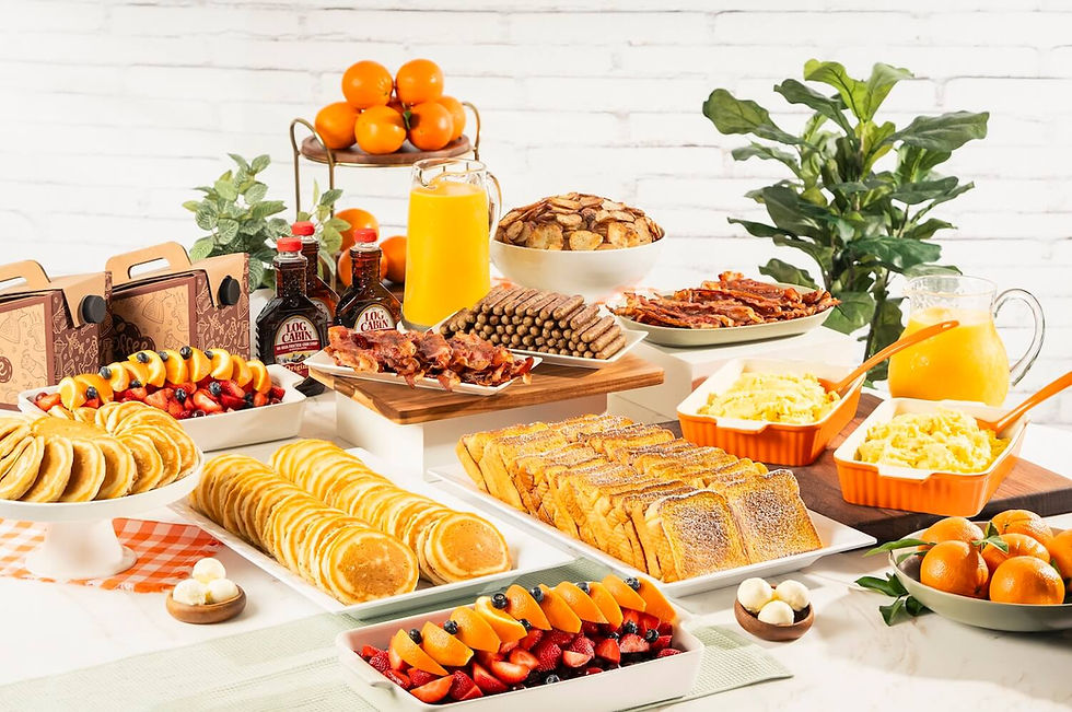 Catering spread of pancakes, French toast, scrambled eggs, bacon, sausage links, fresh fruit trays, oranges, and orange juice pitchers arranged on a white table with to-go boxes