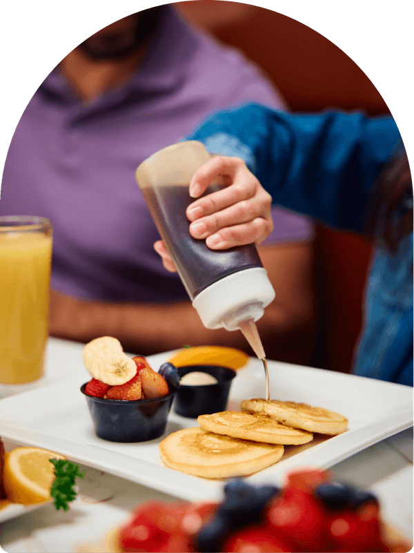 Syrup poured over a short stack of pancakes with fresh fruit and orange juice at Keke's Breakfast Cafe