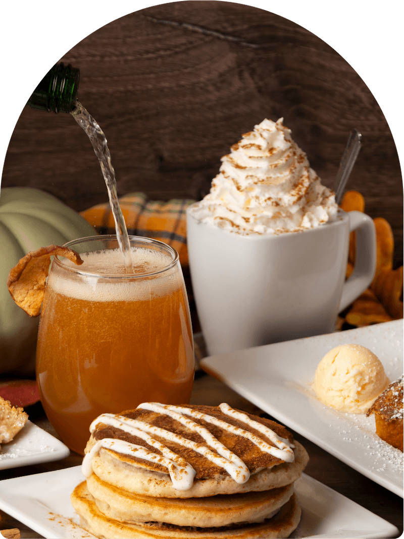 Sparkling wine being poured into an apple cider mimosa beside a pumpkin pie coffee topped with whipped cream, with cinnamon roll pancakes and mini pumpkins on the table