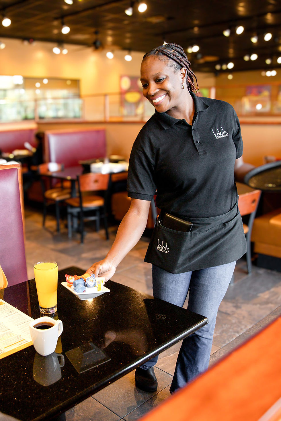 Smiling Keke's Breakfast Cafe server in branded uniform setting jam and butter at a table with coffee and orange juice